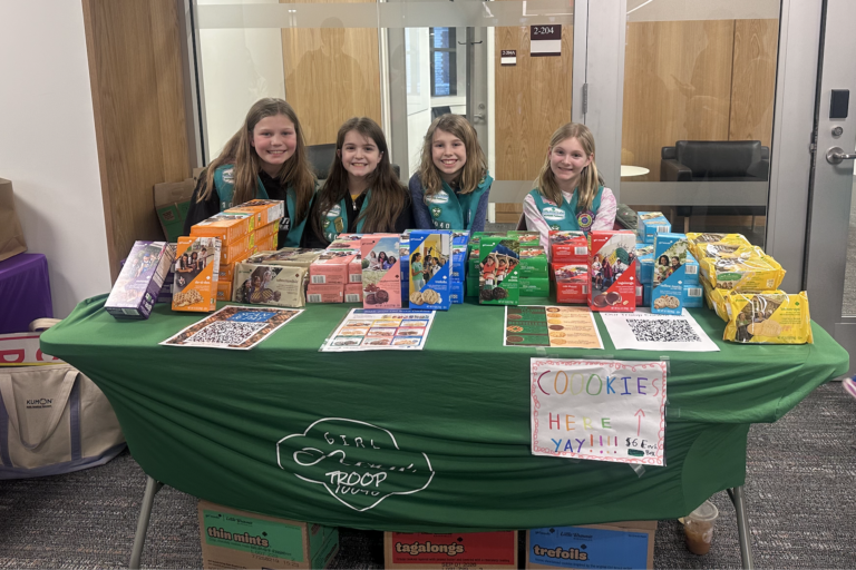 Four young girl scouts behind a green table with a variety of cookies.